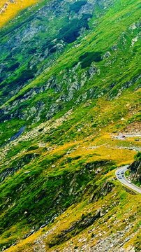 Aerial view of serpentine highway on mountain slope, Romania. Drone shot of winding road, multiple car, green slope and rocky terrain from above. Transfagarash road. Vertical video.