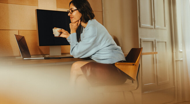 Woman works at desk on laptop while holding a coffee cup in home office