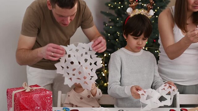 Happy family cut paper snowflakes near Christmas tree, celebrating Christmas with together. New Year's preparations and decorations. Paper crafts. Boy with paper snowflake three people