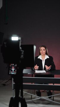 Blurred camera in foreground of studio interview setup. Businesswoman speaks at desk in background with monitor preview and warm lighting. Vertical video.