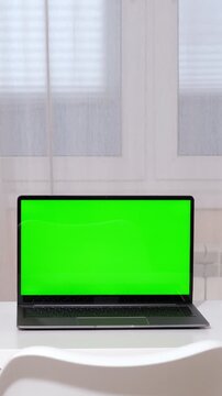 Modern laptop computer with a green chroma key screen sitting on a clean white desk. The bright interior of a home office or living room provides a minimalist backdrop for technology concepts