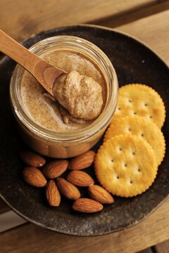 Homemade almond butter in a glass jar with a wooden spoon, served with round crackers and raw almonds on a dark plate, perfect for a healthy snack or cooking inspiration in this stock image