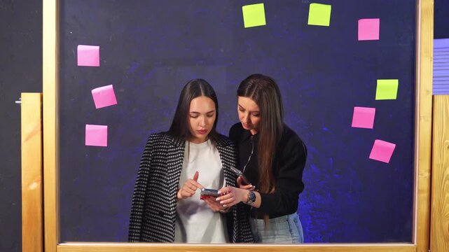 Two colleagues reviewing phone behind glass sticky note board. Overhead pink and green notes surround pair checking data on smartphone.