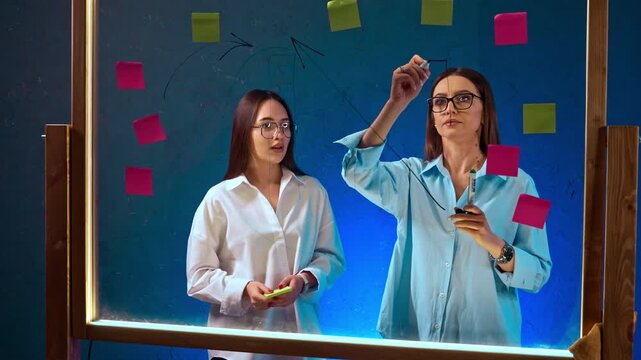 Colleagues sketching arrow diagram on illuminated glass board. Woman draws upward line while partner watches beside blue sticky notes.