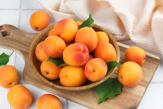 Top view of ripe apricots with green leaves arranged in wooden bowl on on white tile background. Fresh seasonal harvest, summer fruit, healthy eating, dessert, food ingredient