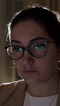 Close up of a professional woman with glasses concentrating on her computer screen in a dimly lit environment, her face illuminated by the monitor as she works on a digital project