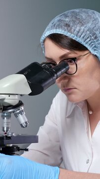 Concentrated female microbiologist in a sterile cap and gloves, meticulously analyzing a sample using a laboratory microscope, engaged in scientific research and medical development