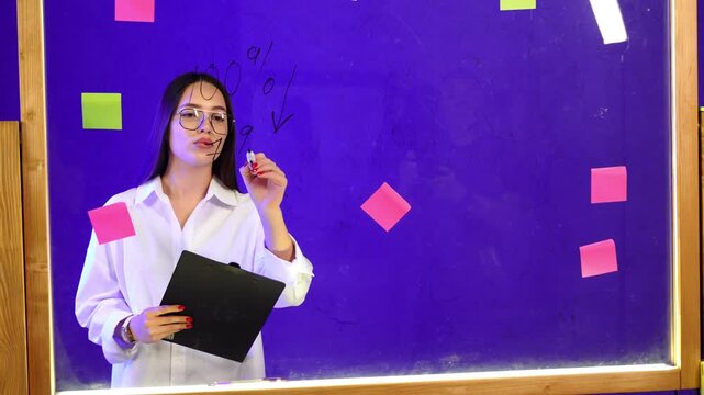 Businesswoman drawing declining percentage chart on purple glass. Analyst marks downward arrow while holding clipboard against bright backdrop.