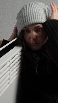 Freezing sad woman wearing a warm hat and sweater suffering from the cold, trying to get warm near an electric heater during the central heating shutdown and energy crisis