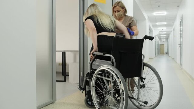 Young woman with disability sits down into wheelchair with medic support in clinic. Nurse cares of female patient during hospital procedures