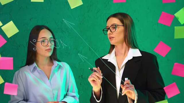 Girls gesturing during discussion at green glass board. Woman draws arrow line while partner listens beside scattered sticky notes.