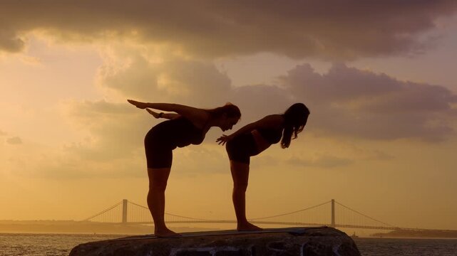 Women performing standing half fold stretch outdoors. Two female silhouettes extend arms backward on concrete pier with bridge in background, New York.