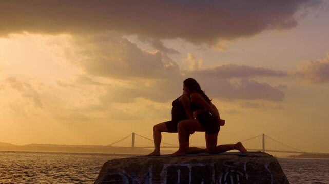 Two women holding bound low lunge stance at dusk. Female silhouettes interlock hands behind back on stone pier near Verrazzano Narrows Bridge.