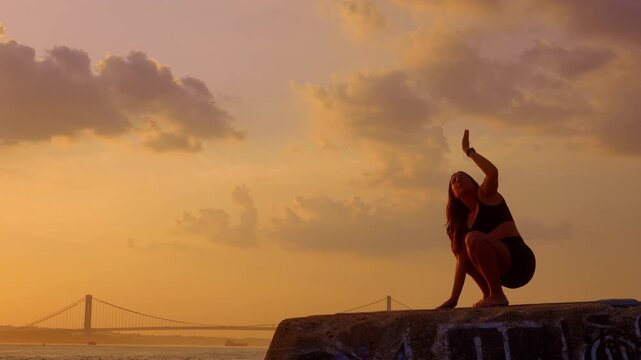 Woman executing deep squat yoga pose at sunset near coastal bridge. Side view silhouette of female practitioner on stone block by water. Yellow sky and clouds in background.