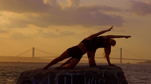 Two women holding lateral plank pose on coastal pier. Female silhouettes execute side body stretch on stone surface against sunset sky in New York.