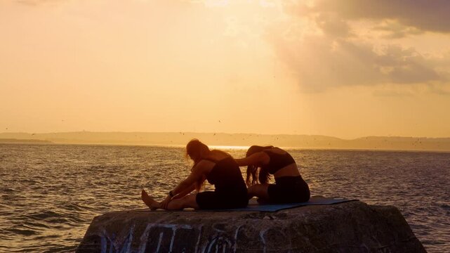 Two women performing seated forward stretch near water. Female athletic silhouettes bend over legs on stone breakwater during golden evening light in New York.