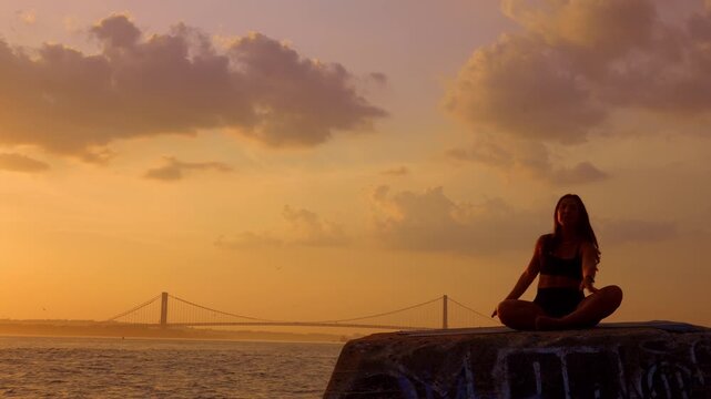 Woman sitting in meditation pose during golden hour near New York harbor.. Female figure in cross legged posture on stone block. Warm evening lighting, clouds and large suspension bridge behind.