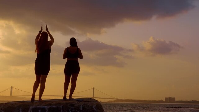 Women standing in mountain pose facing sunset water. Two female figures hold prayer hands position on waterfront block with Verrazzano Narrows Bridge behind.