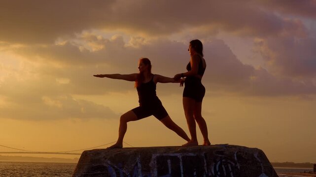 Woman performing warrior pose with instructor guidance by the water. Silhouetted duo executing yoga posture on stone pier. Golden dusk light and suspension bridge in background.