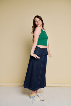 Teenage girl poses with joy in a green tank top and denim skirt against beige backdrop