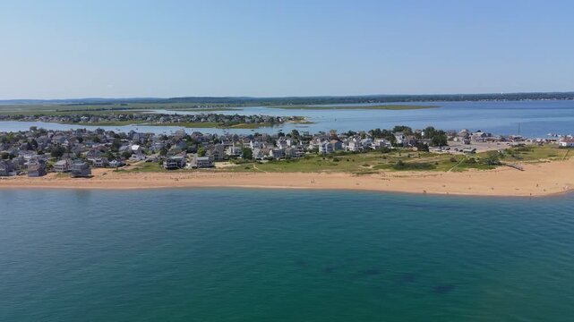 Rows of closely packed beachfront homes line narrow sandy peninsula between two wide channels of calm blue water, salt marsh grass frames coastal neighborhood in USA. Aerial drone wide shot