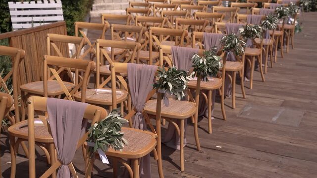 Chairs are set in neat rows for a wedding ceremony. Each chair has a decoration with fabric and green leaves. The setting is an outdoor place with wooden flooring.