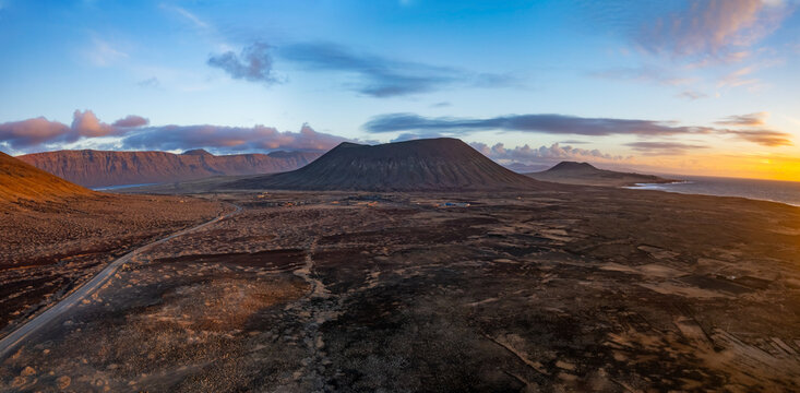 The scene captures Lanzarote and La Graciosa Canary Island at sunset, showcasing volcanic hills and coastline. The sun sets over the ocean, enhancing the landscape features.