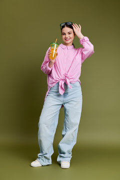 Stylish teen stands confidently on olive green backdrop, holding a refreshing drink