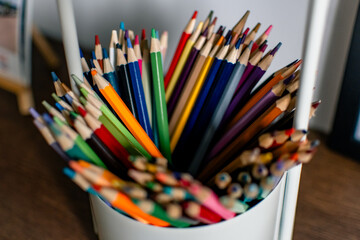 A white cylindrical pencil organizer with a tall, arched handle, filled to the brim with a multitude of sharpened colored pencils. The pencils are arranged haphazardly, showcasing a variety of vibrant © Yulia