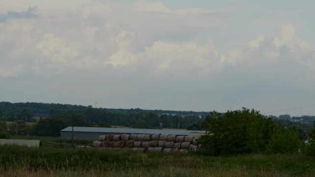 A stack of hay bales near farm buildings under clouds.