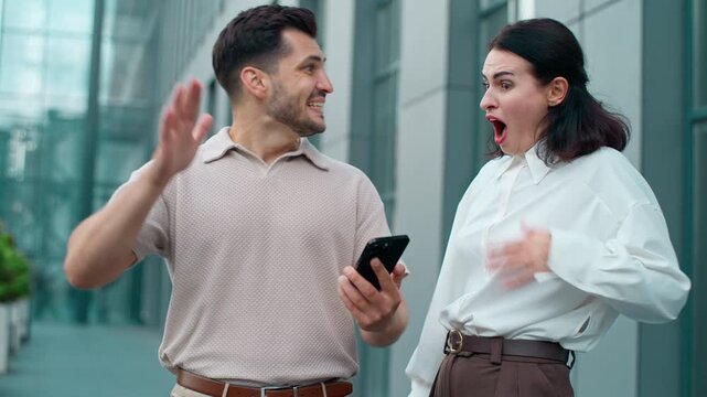 Two excited happy shocked Caucasian business colleagues winning with mobile phone giving high five in city. Positive amazed man with woman businessman with businesswoman win with smartphone celebrate