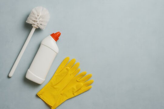 Toilet brush, cleaning solution, and yellow rubber gloves arranged on a light gray surface for bathroom cleaning.