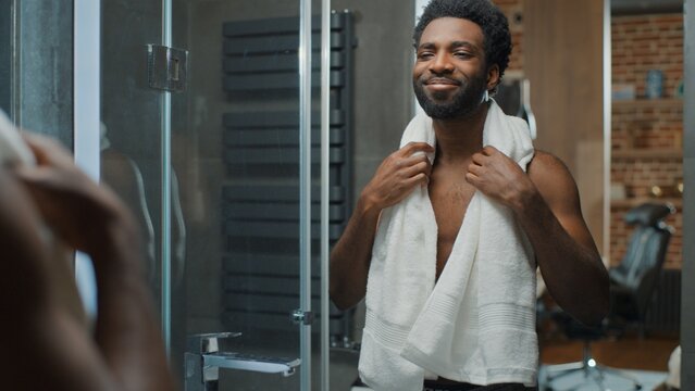 Handsome African American man smile at mirror reflection touching soft beard hair after gel balm masculine cosmetic in bathroom at home smiling male after hygiene in bath guy touch facial smooth skin