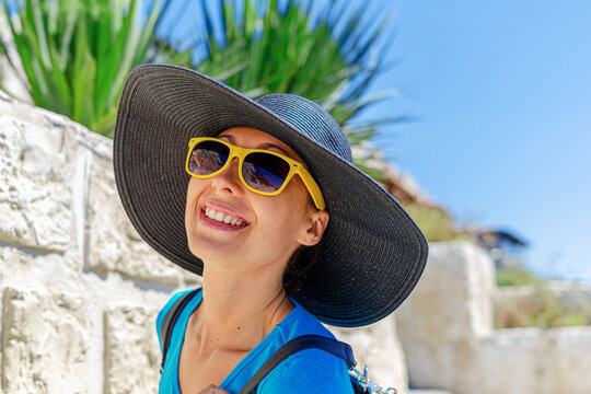 A happy woman wearing a wide brim straw hat and bright yellow sunglasses smiles in the sunshine during a summer vacation.
