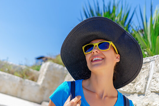 A smiling woman wearing a large black sunhat and bright yellow sunglasses enjoys a sunny outdoor vacation day.