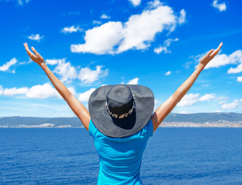 A happy woman in a sunhat stands with her arms raised while looking at the sea under a bright blue sky.
