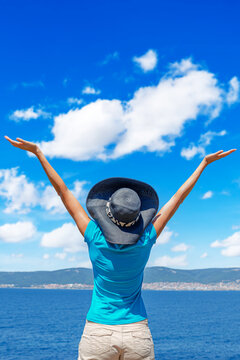 A happy woman stands with her arms raised wide towards the bright blue sky over the sea.