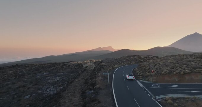 Car journey in Tenerife, white cabriolet moving along a winding asphalt road during sunset. Travel to Canary Islands, with the majestic Teide volcano nature landscape. Aerial tracking drone flight