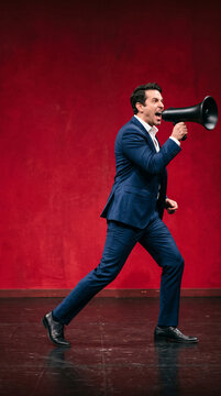 Man in blue suit shouting into a megaphone on a red stage, symbolizing announcement, power, and communication, suitable for business or public speaking.