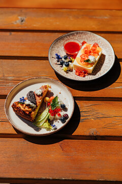 Two elegant plated desserts with cake, berries, cream, citrus, jelly cubes, edible flowers, and bright fruit sauce arranged on a sunlit wooden table