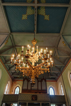 Elaborate golden chandelier with glowing candle-style lights beneath a wooden vaulted ceiling, framed by arched church windows and a pipe organ