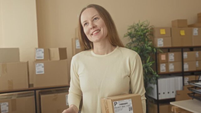 Woman holding parcel and smartphone, hand presenting package amid stacked boxes and shelving in building; small business calm efficiency.