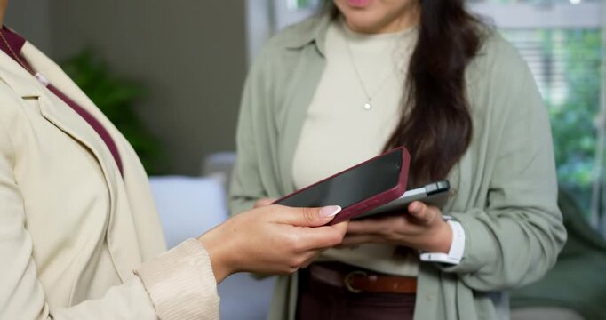 Diverse female coworkers comparing devices, left offering phone and right checking tablet on sofa