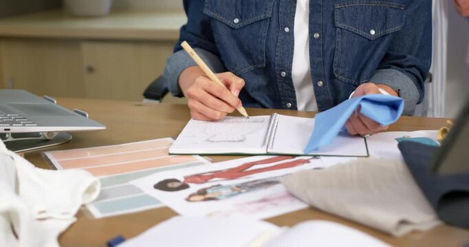 Picking up blue fabric swatch, designer is sketching garment details and judging drape for fit