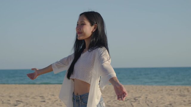 Happy free woman enjoying nature outdoors, open arms feel fresh air, beautiful Asian girl with long hair smiling on sandy ocean beach under blue sky, finding mental health freedom.