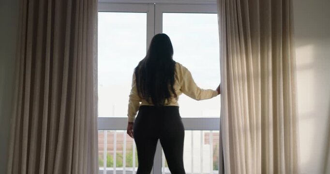 Facing curtains, woman is reaching up, pulling beige curtains aside at window to look outside
