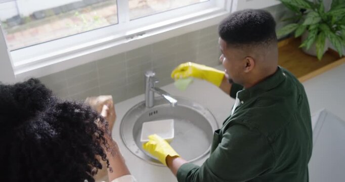 African American couple washing dishes after dinner at kitchen sink, man scrubbing in yellow gloves