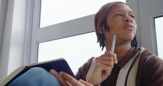 Closeup hands urging African-American man writing journal at window tapping pen musing, copy space