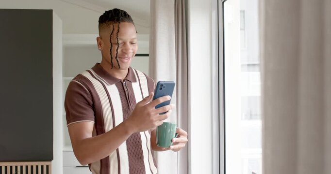 African American man brown polo by large window, glancing at blue phone while holding green mug