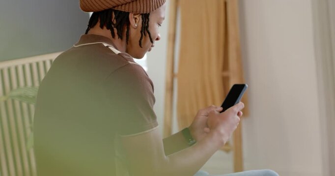 African American man scrolling smartphone on bed edge by window, wearing beanie and smartwatch